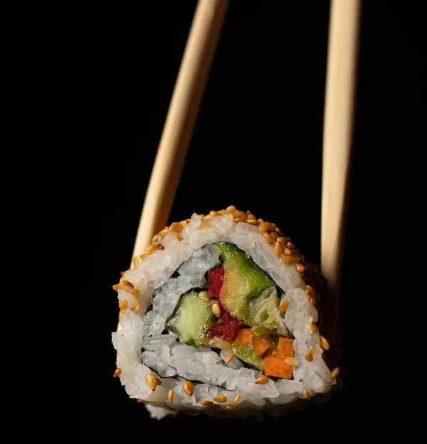 A sushi roll filled with avocado, cucumber and carrot, held up in wooden chopsticks